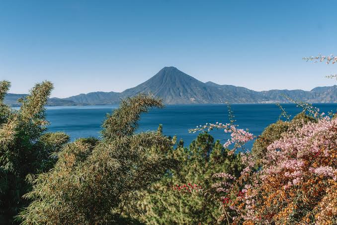 Volcano rising above a blue lake beyond green trees and flowers